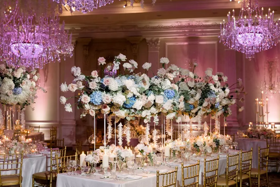 A long table set beneath a towering arrangement of white roses and blue hydrangeas, chandeliers above.