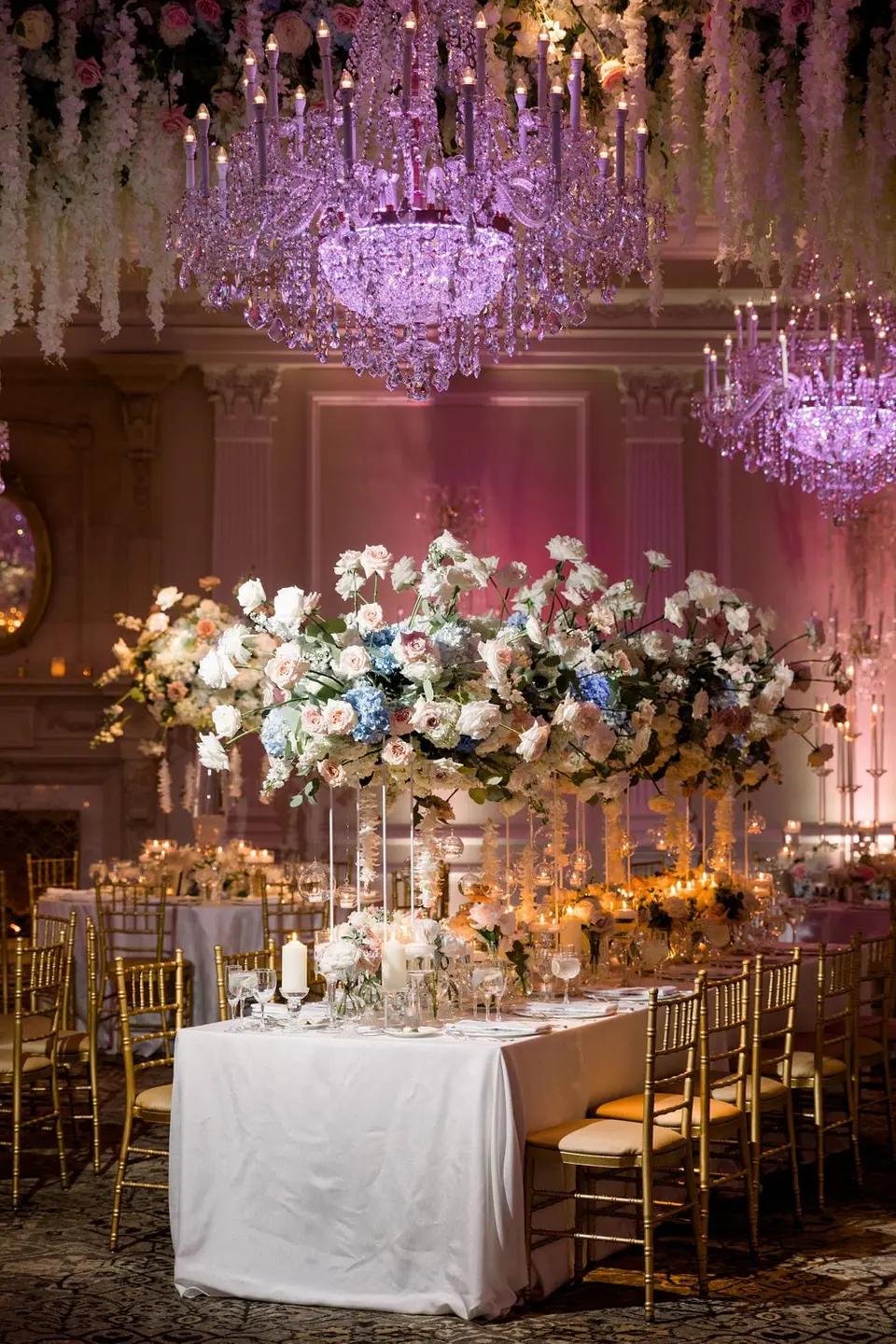 A long table beneath a tall arrangement of white roses and blue hydrangeas, hanging white flowers overhead.