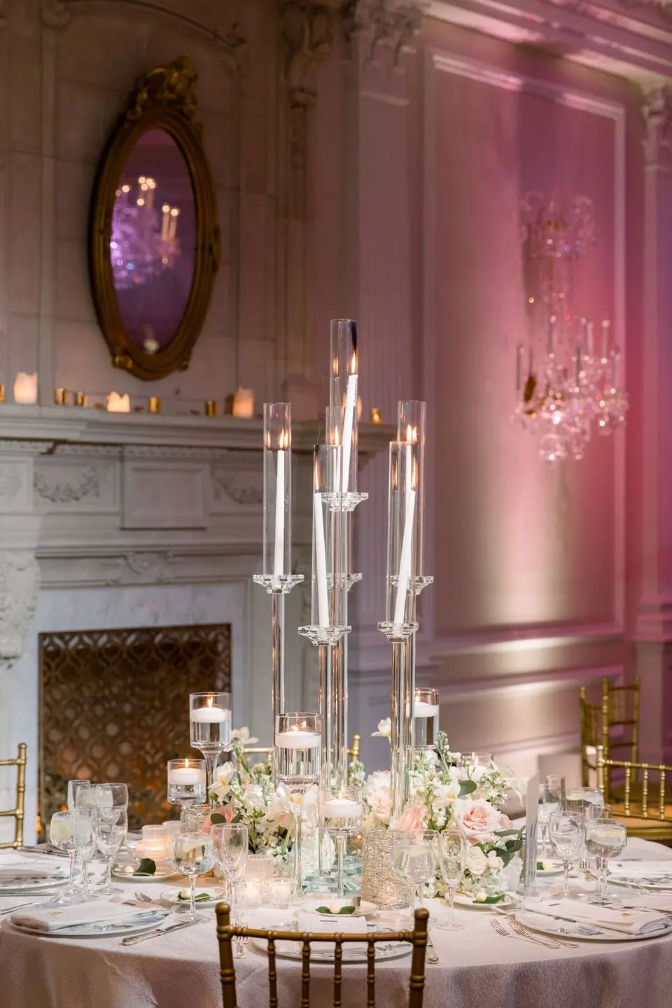 Tall glass candle holders above a round table of white flowers, beside a marble fireplace.