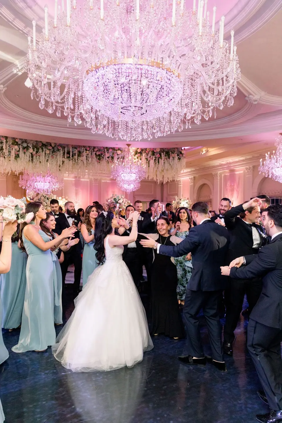 The bride and groom dance in a circle of guests beneath a crystal chandelier.
