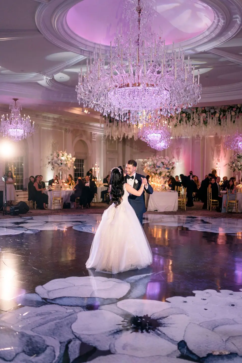 The bride and groom dance beneath a great chandelier, the floor painted with white blooms.