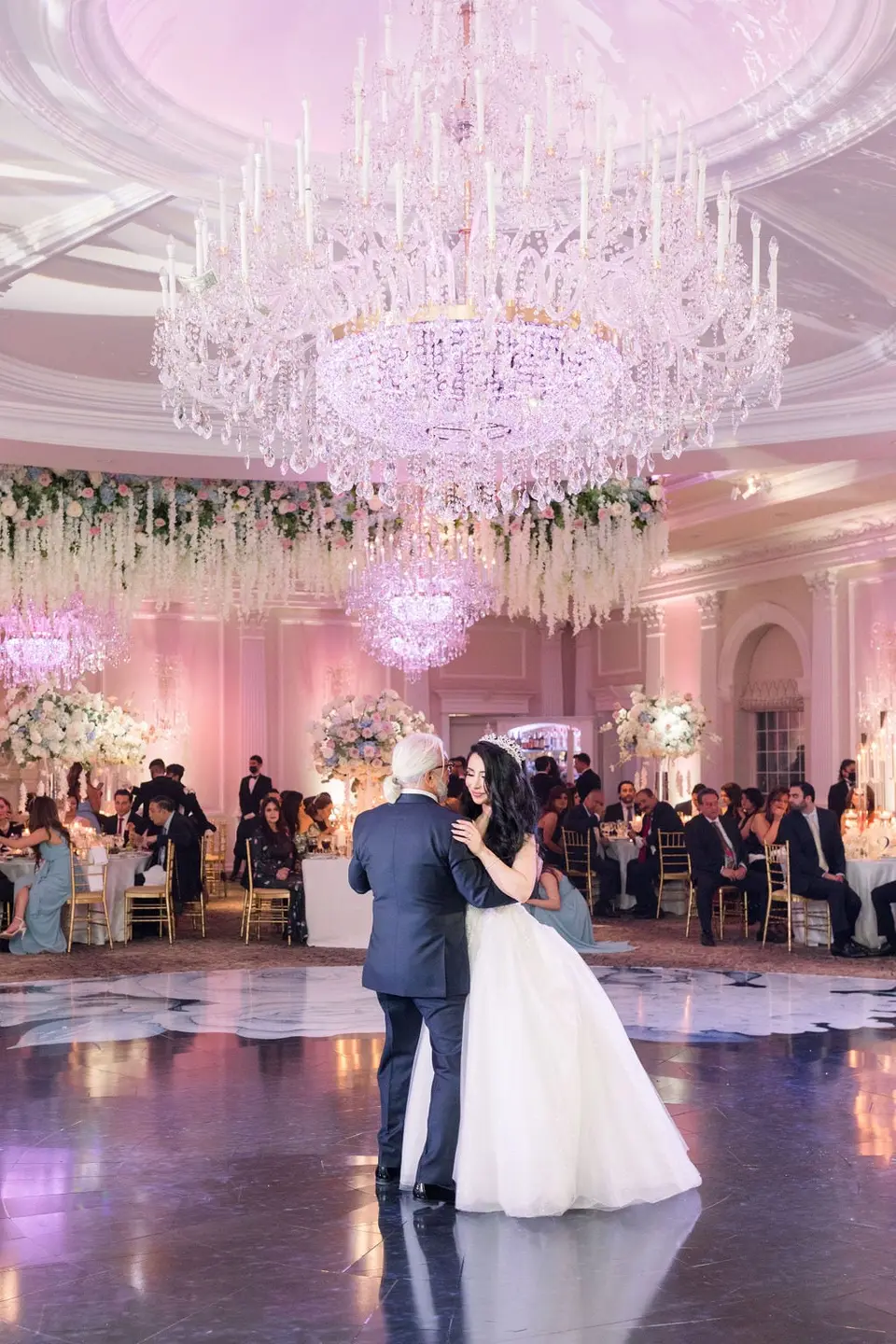 The bride dances with her father beneath a great crystal chandelier.