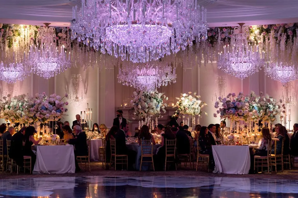The ballroom full of guests at dinner beneath crystal chandeliers and hanging white flowers.