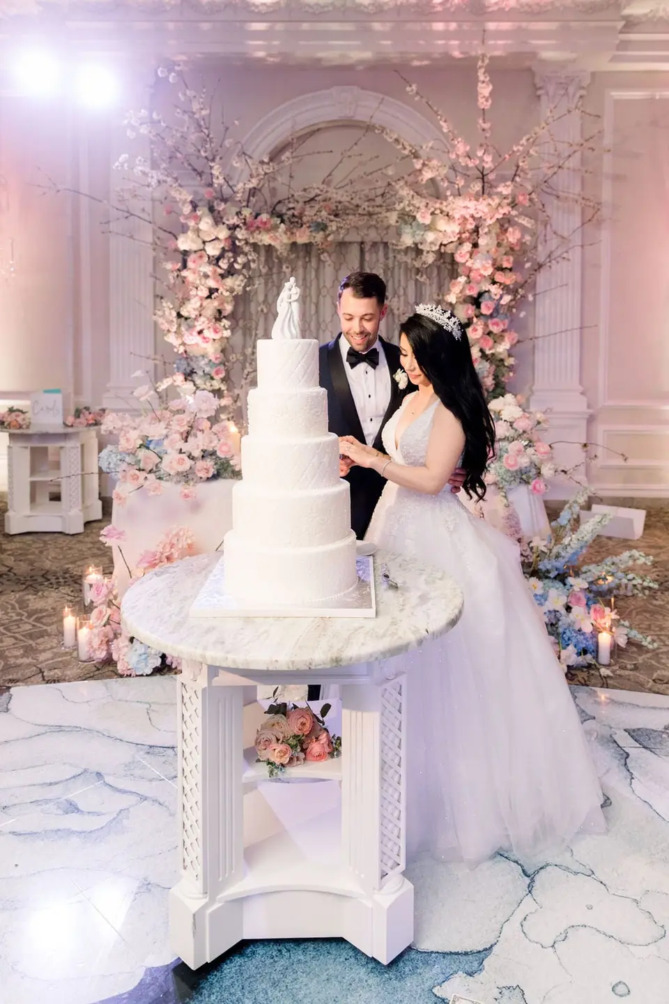 The bride and groom beside a white four-tier cake before the flowered arch.