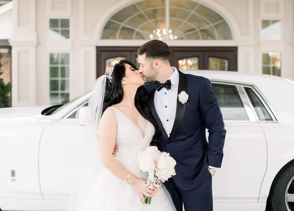 The couple kiss beside a white Rolls-Royce at the entrance of The Rockleigh.