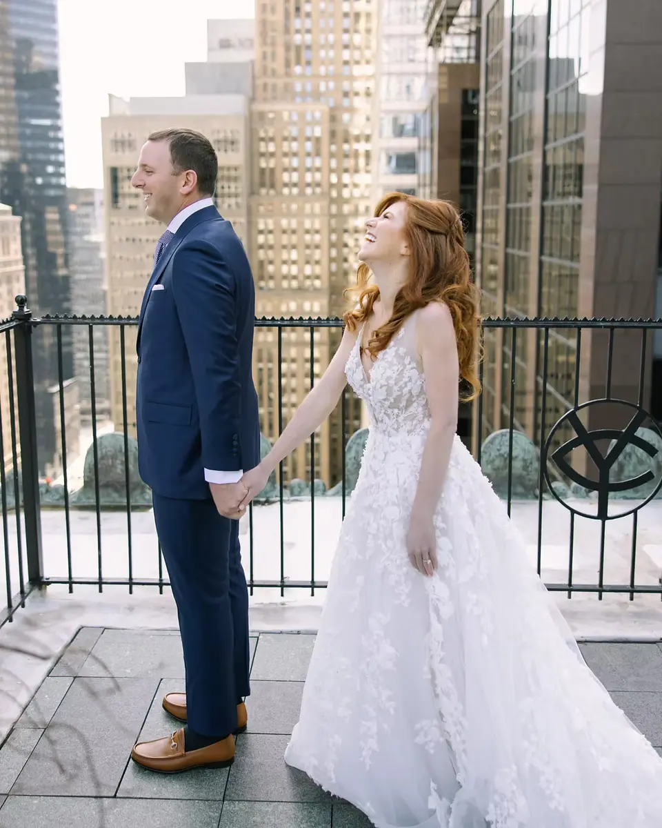 The couple face each other on a rooftop terrace above Midtown, a black railing and glass towers behind them.