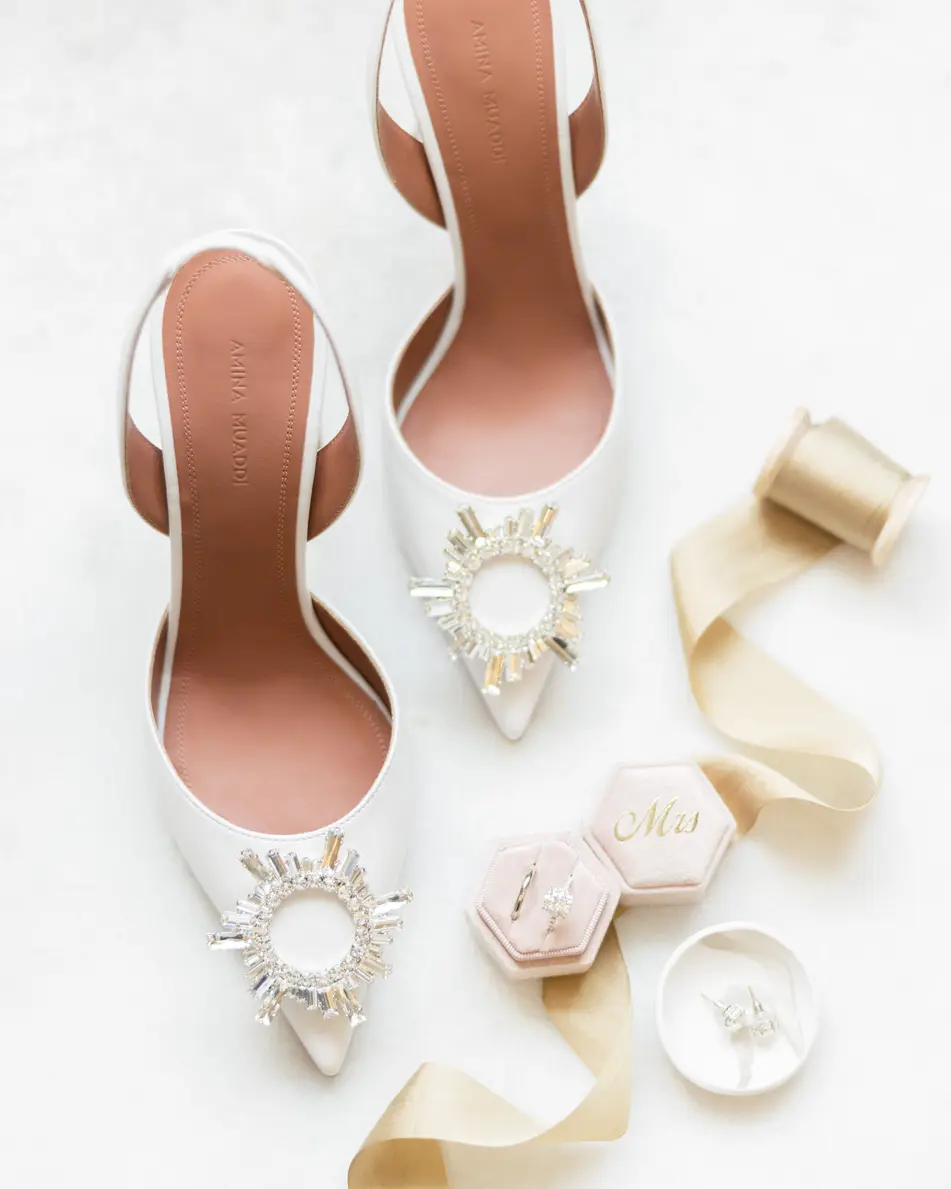 The bride's white shoes with jewelled buckles, laid on white with silk ribbon and a ring box.