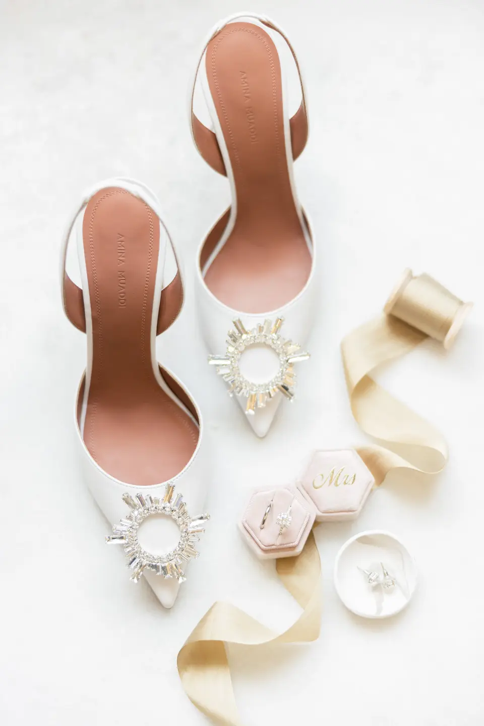 The bride's white shoes with jewelled buckles, laid on white with silk ribbon and a ring box.