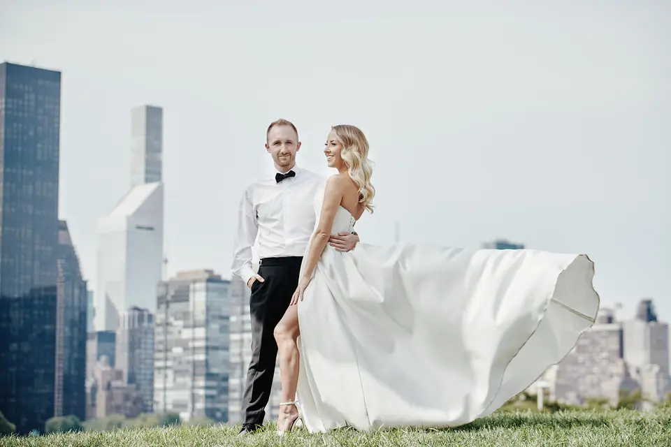 The wind lifts the bride's skirt as she stands with the groom on the grass above the river, the skyline behind.