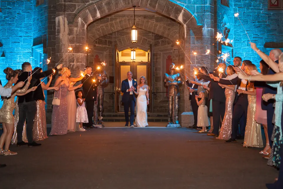 Guests hold sparklers in two lines outside the castle doors, lit in blue, as the couple walk out between them.