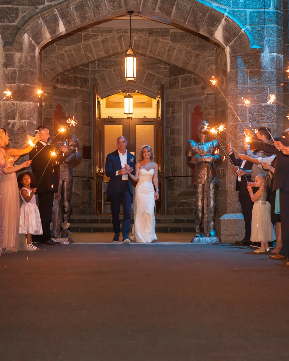 Guests hold sparklers in two lines outside the castle doors, lit in blue, as the couple walk out between them.