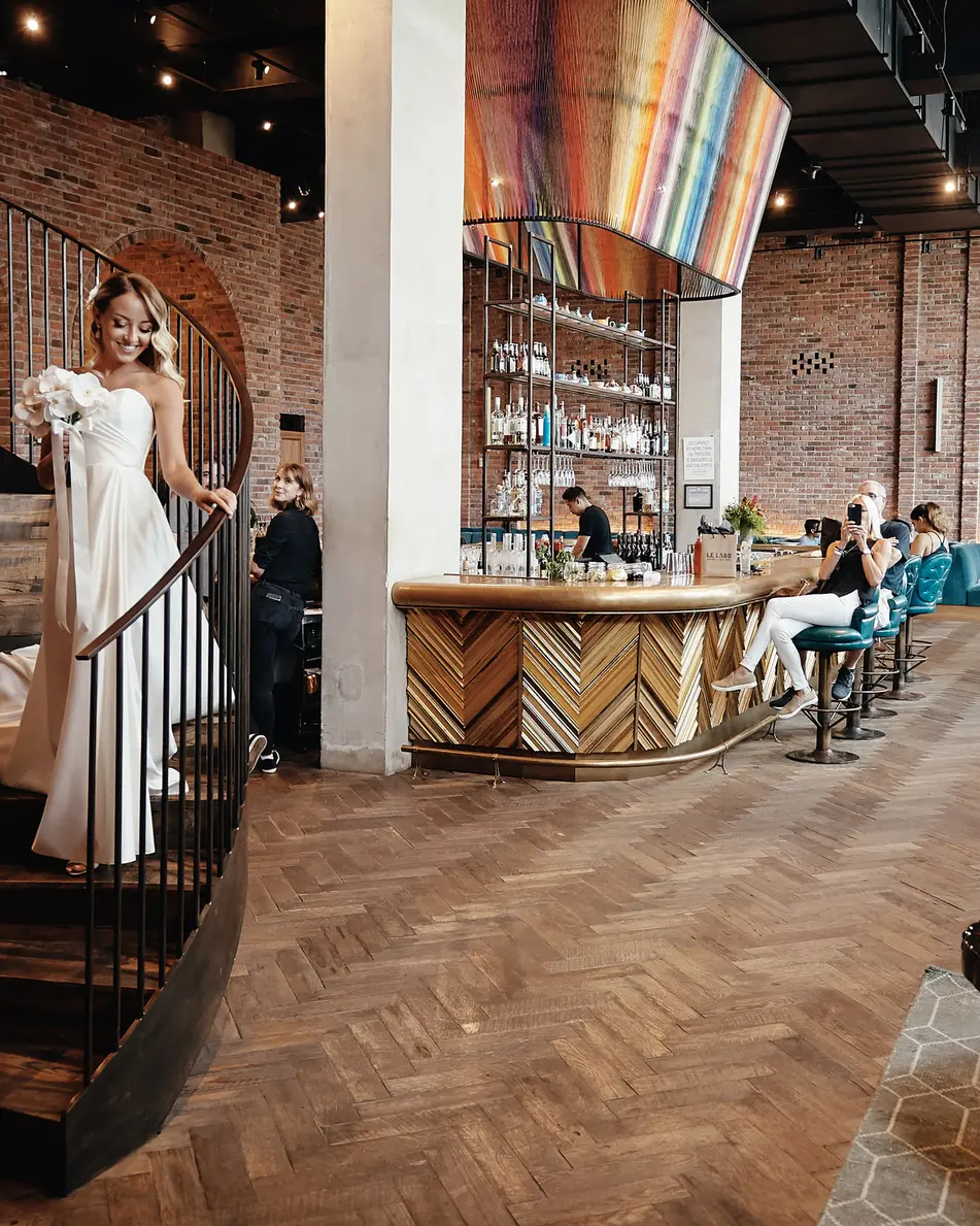 The bride comes down a spiral staircase into a lobby of brick walls and warm lights, the groom waiting below.