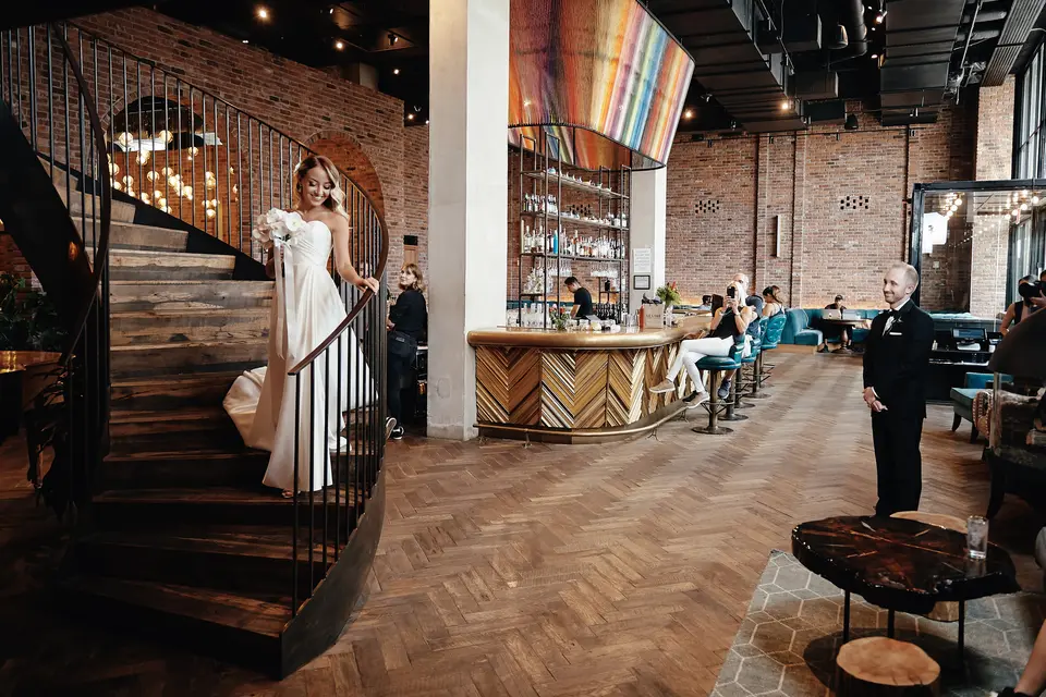 The bride comes down a spiral staircase into a lobby of brick walls and warm lights, the groom waiting below.