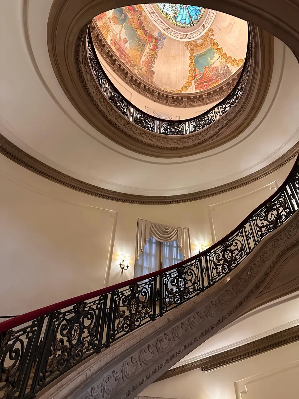 Looking up the spiral marble staircase of the Burden Kahn Mansions, its black iron balustrade curving beneath a painted dome.