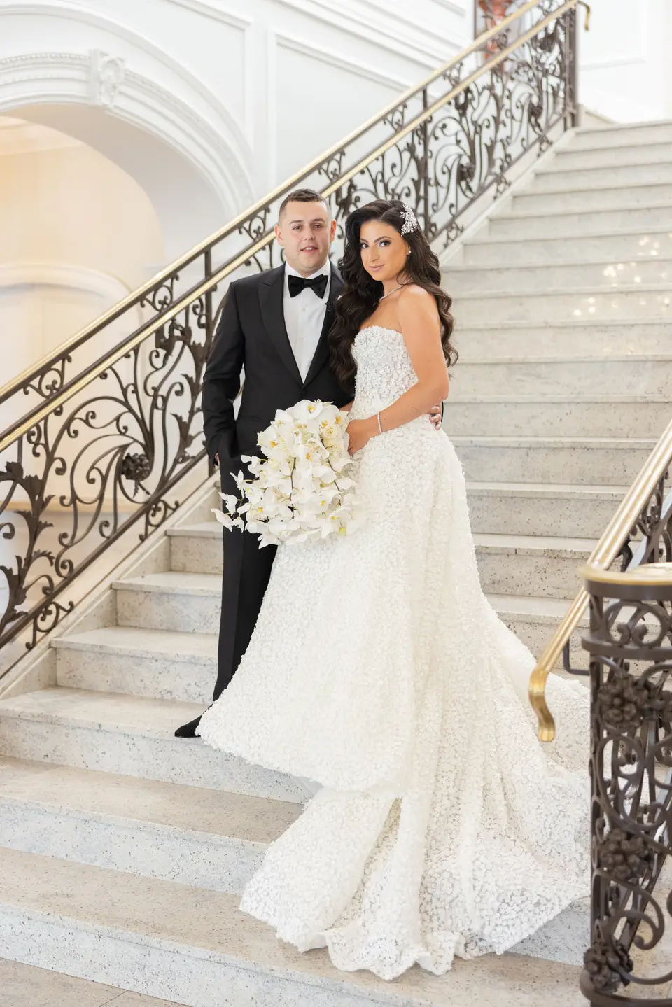 The couple on the white marble staircase at The Rockleigh, its wrought iron balustrade curving above them.