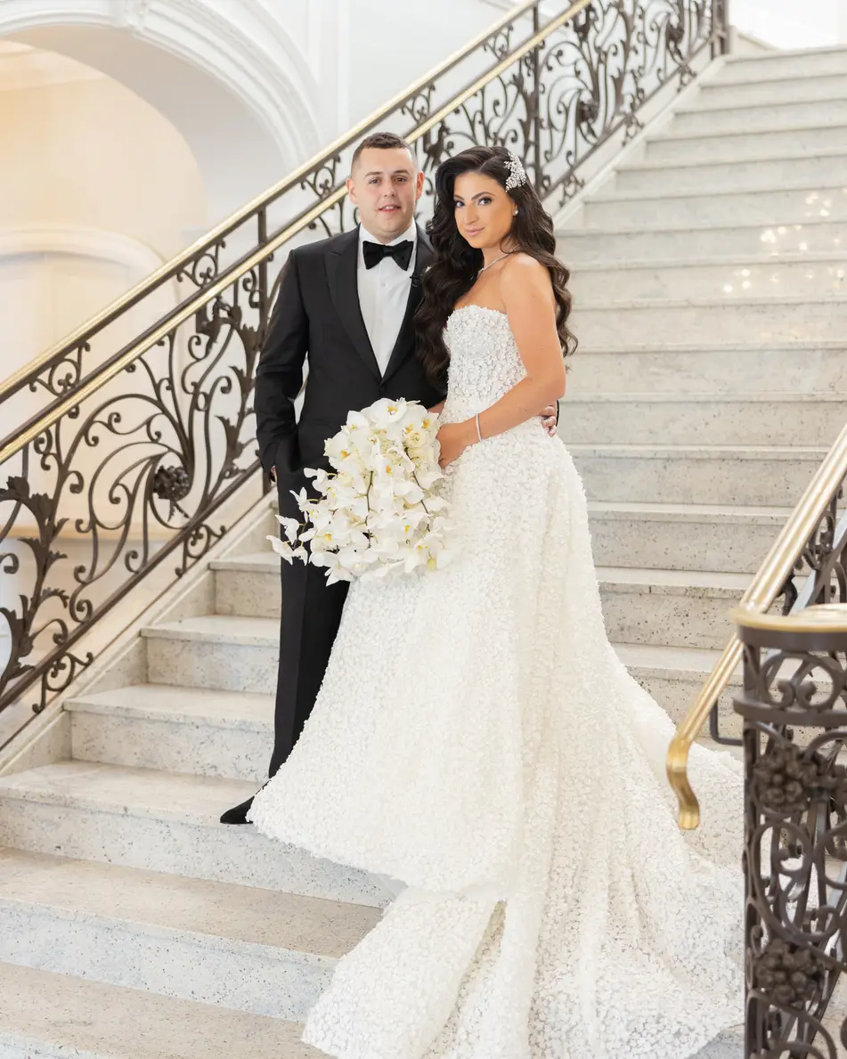 The couple on a white marble staircase with a wrought iron balustrade, the groom a step above, the bride's train across the stairs.