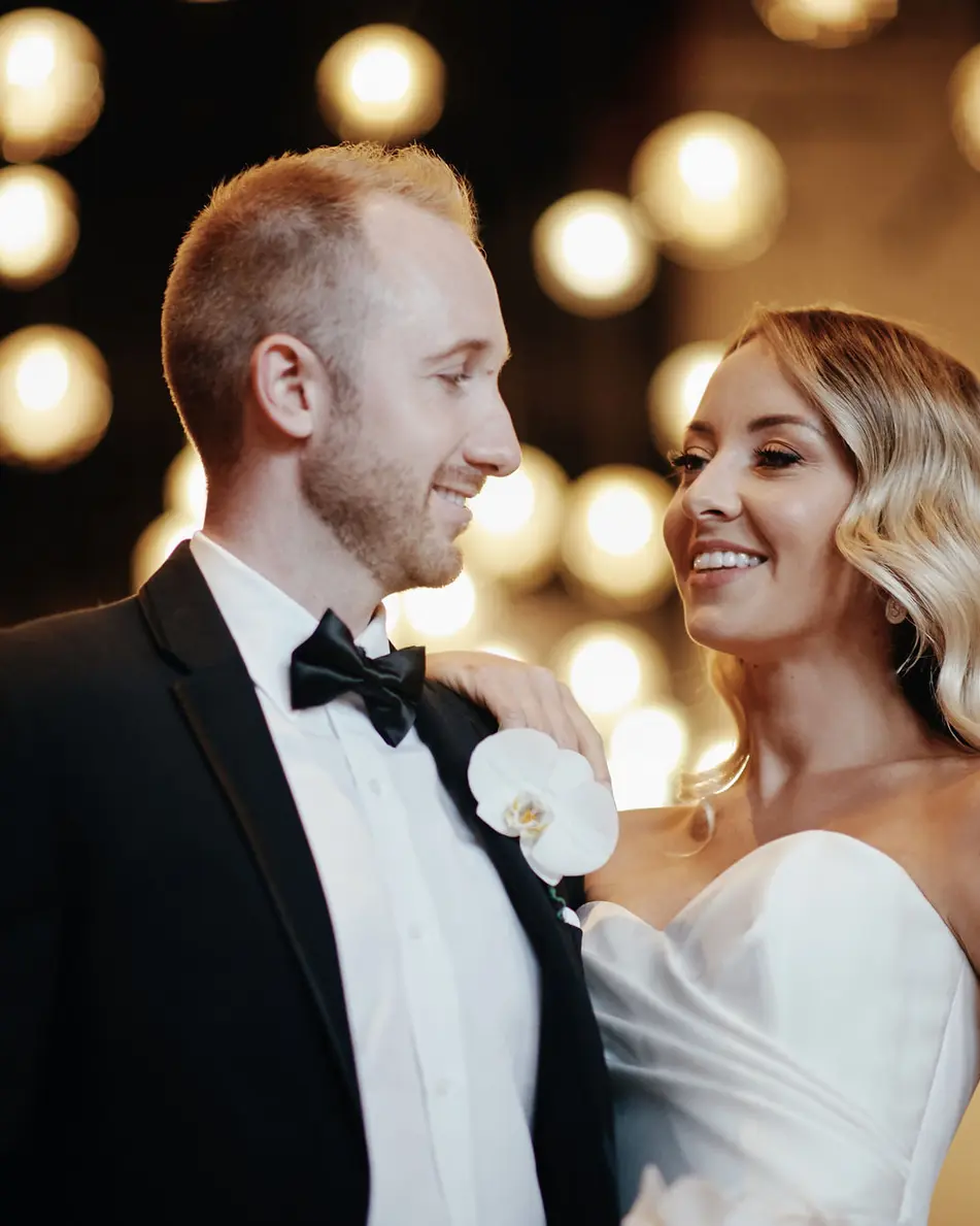 The couple smile at each other in front of a wall of glowing bulbs.
