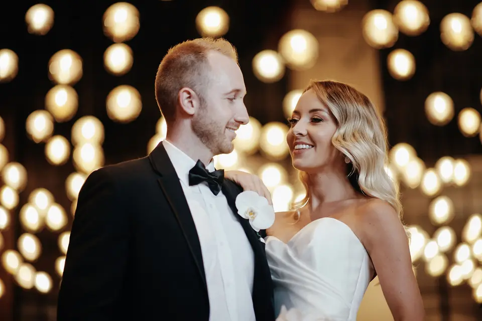 The couple smile at each other in front of a wall of glowing bulbs.