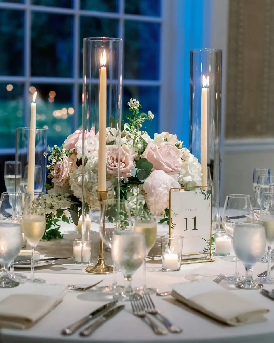 A dinner table with tall candles, blush roses and glassware, deep blue dusk in the windows behind.