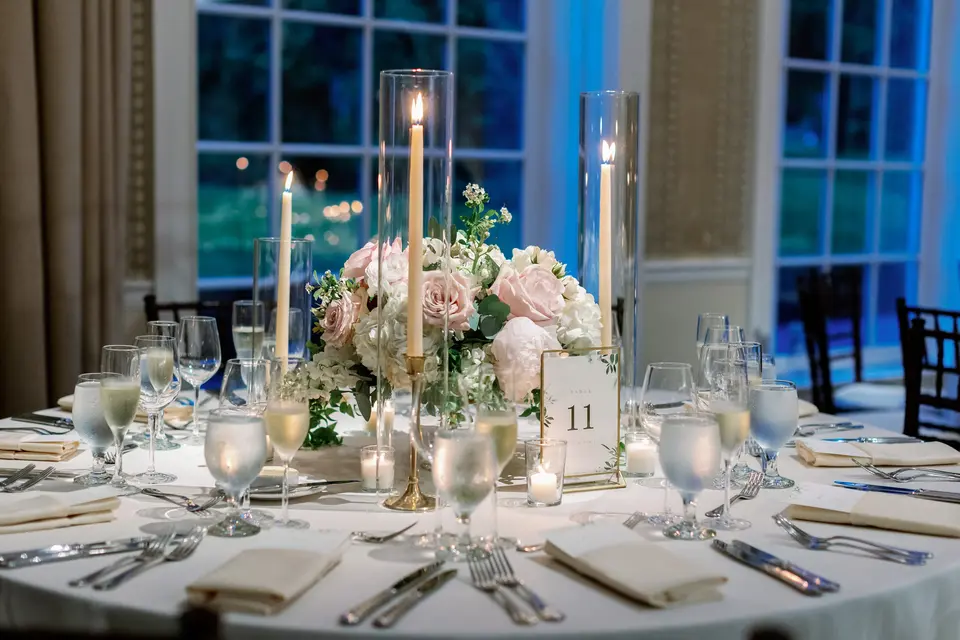 A dinner table with tall candles, blush roses and glassware, deep blue dusk in the windows behind.