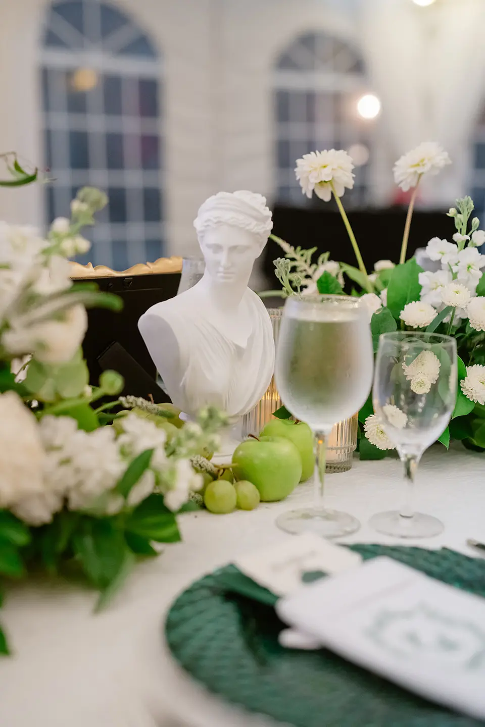 A table with a small white classical bust, white flowers, greenery and green limes.