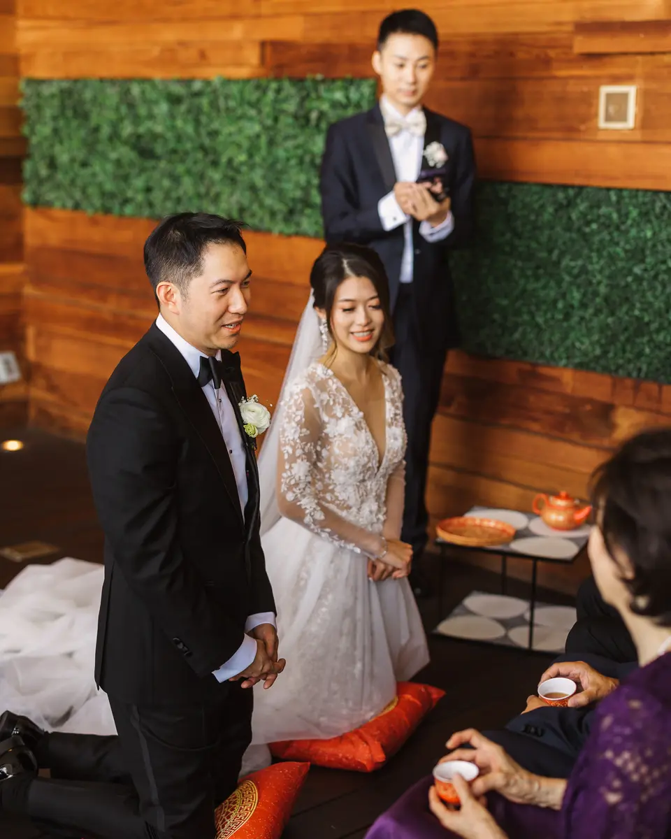 The couple kneel on orange cushions before an elder, a tray of tea cups between them.