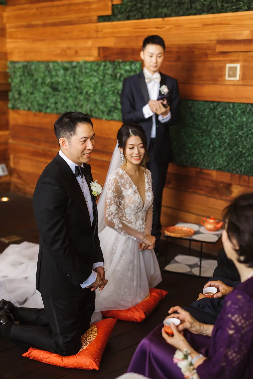 The couple kneel on orange cushions before an elder, a tray of tea cups between them.