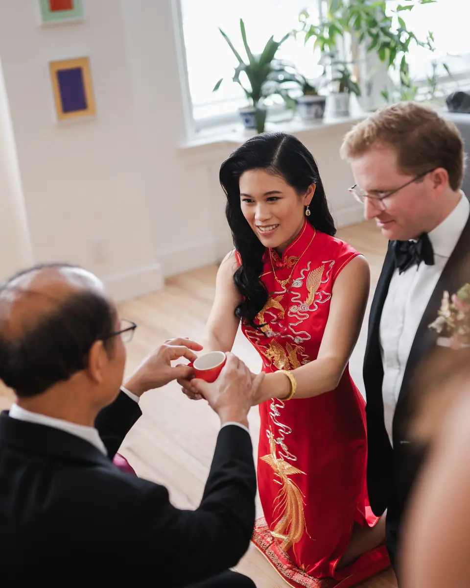 The bride, in a red qipao embroidered in gold, offers a cup of tea to a seated elder, the groom in black tie beside her.