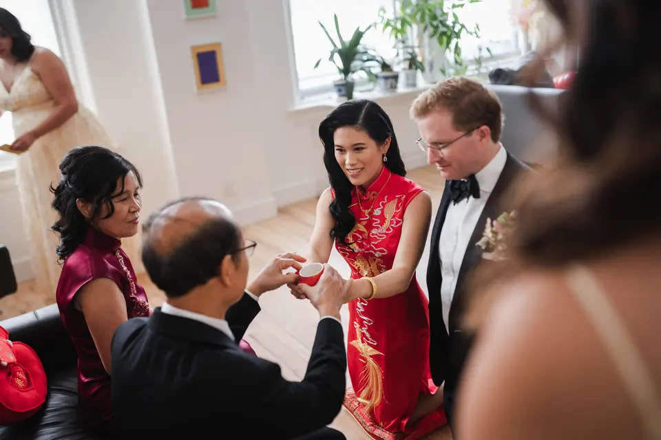 The bride, in a red qipao embroidered in gold, offers a cup of tea to a seated elder, the groom in black tie beside her.