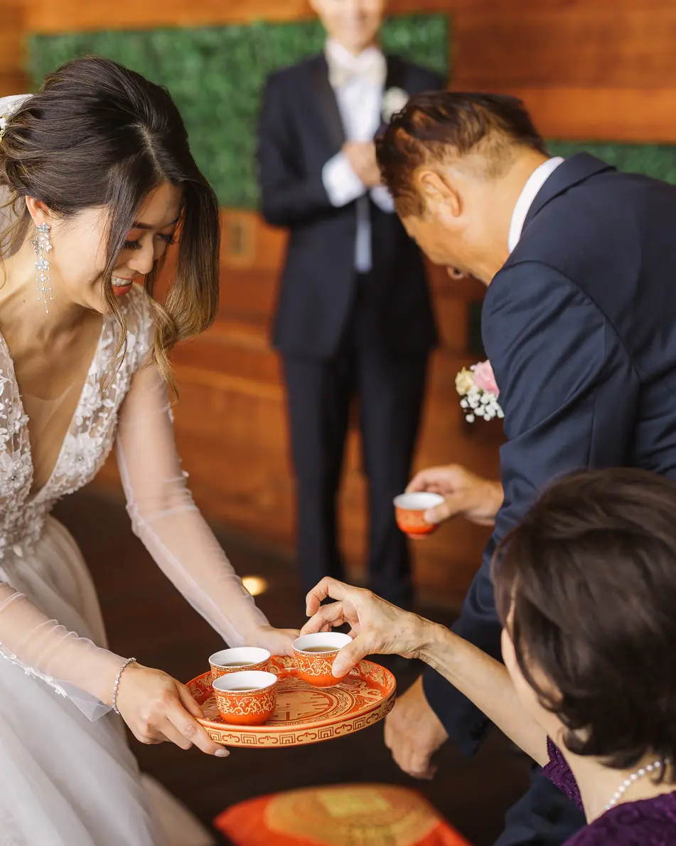 The bride, bending low, offers a tray of tea cups to an elder while an older man in a navy suit leans in beside her.