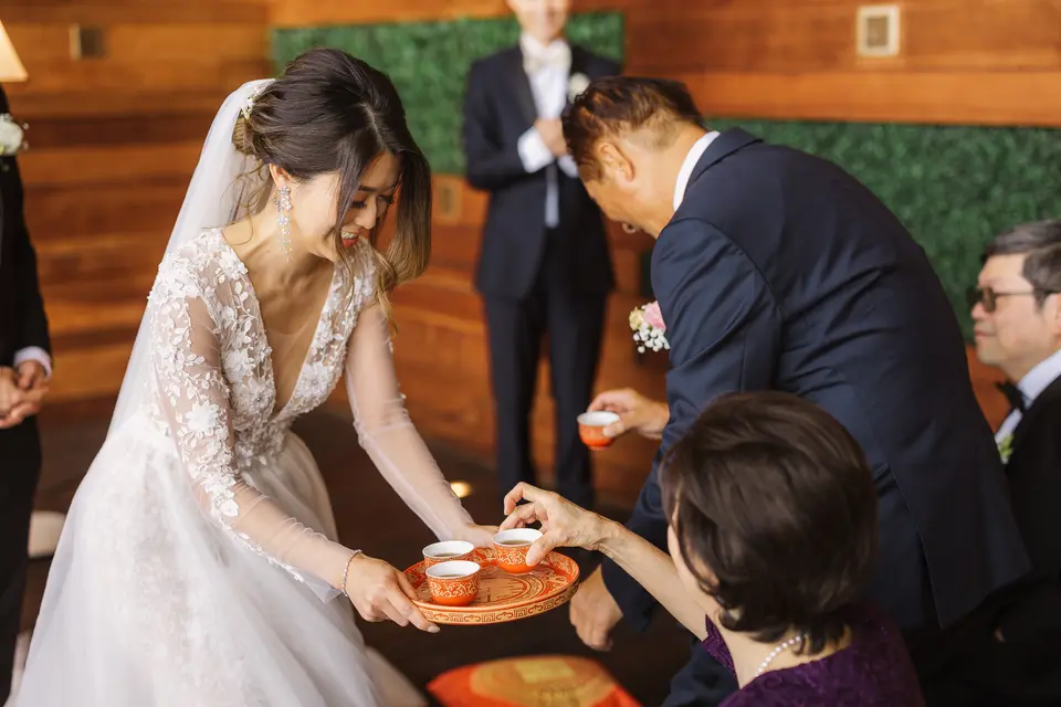 The bride, bending low, offers a tray of tea cups to an elder while an older man in a navy suit leans in beside her.