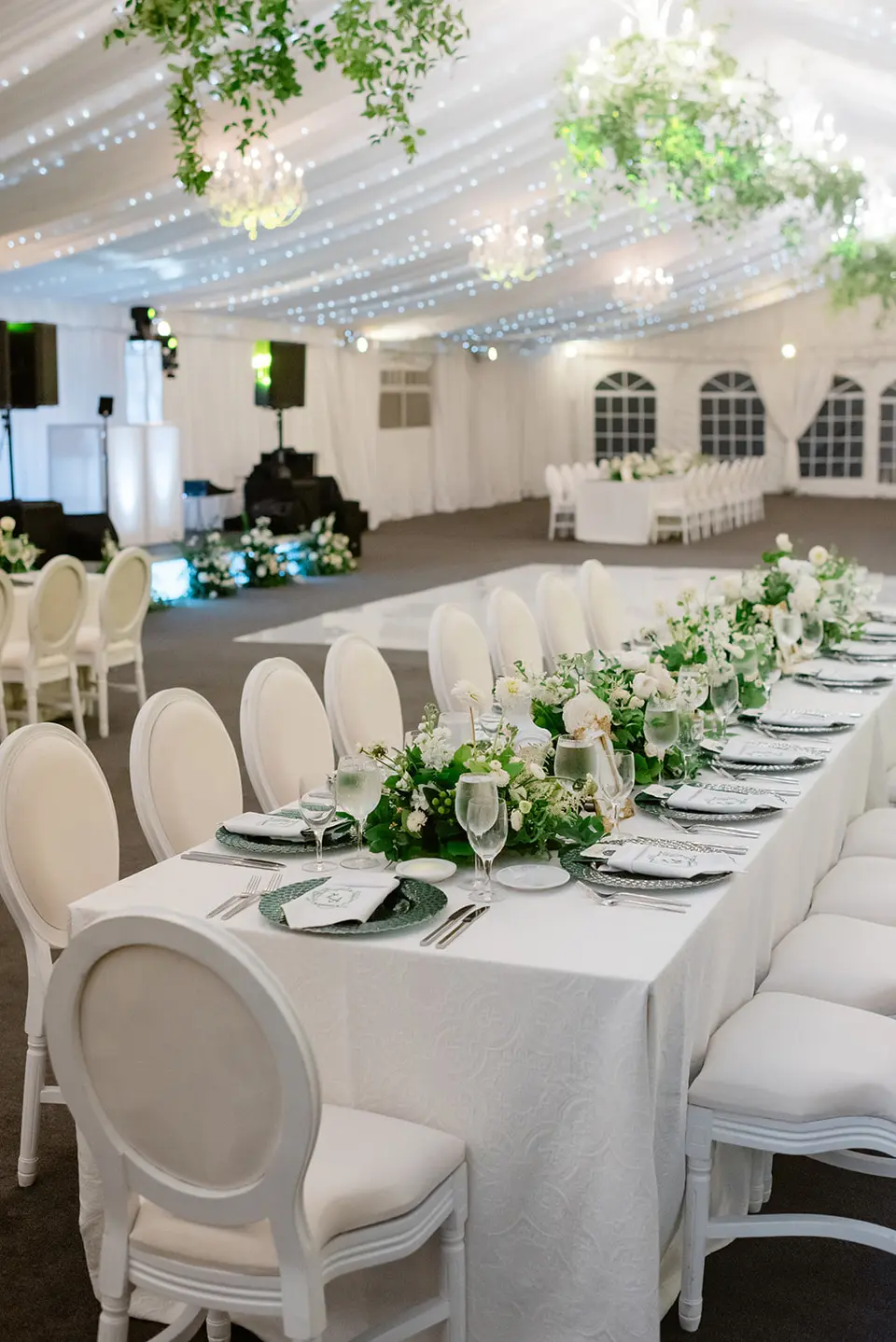 A long table in white linen and white chairs, greenery running down its length, under the draped tent ceiling.