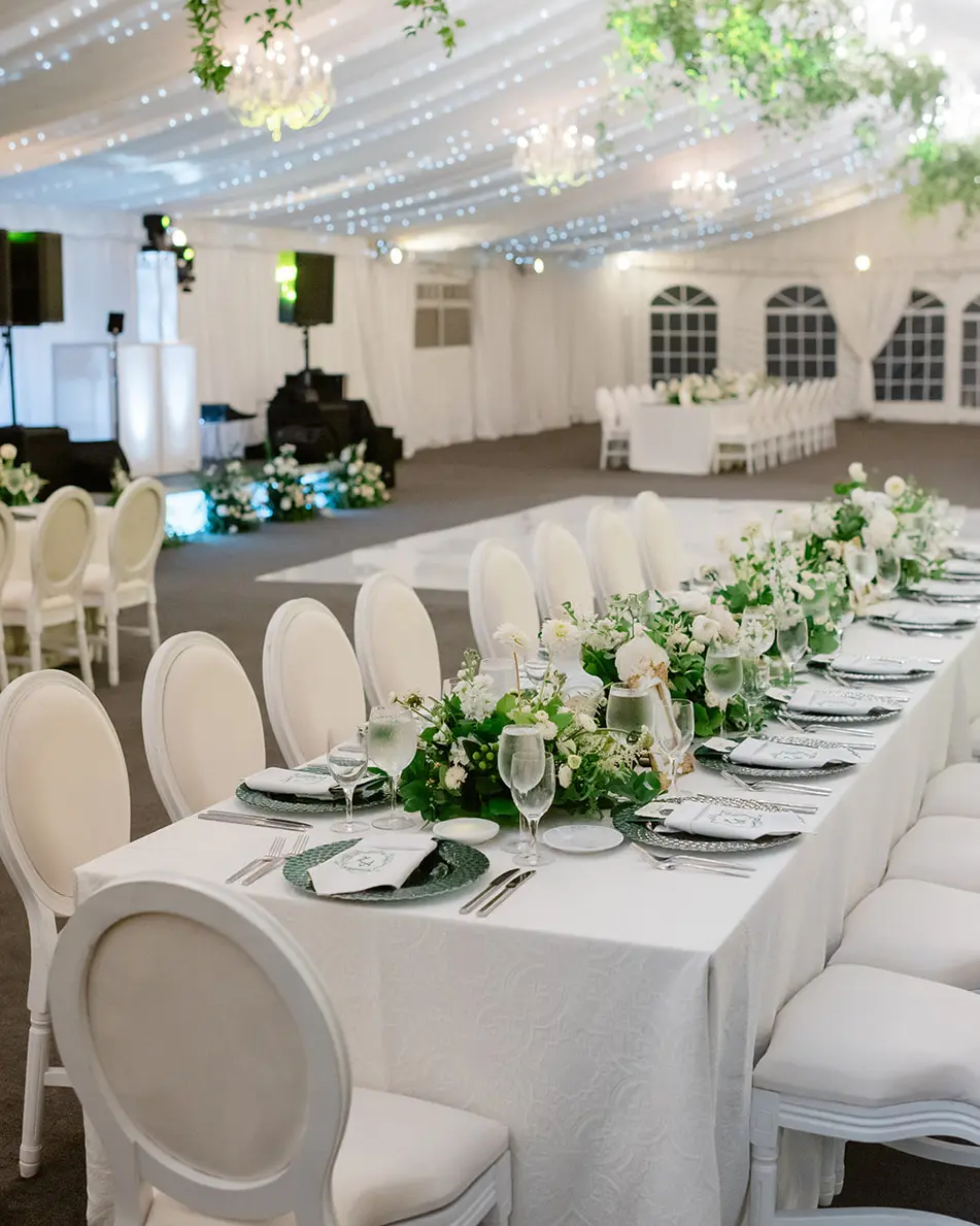 A long table in white linen and white chairs, greenery running down its length, under the draped tent ceiling.