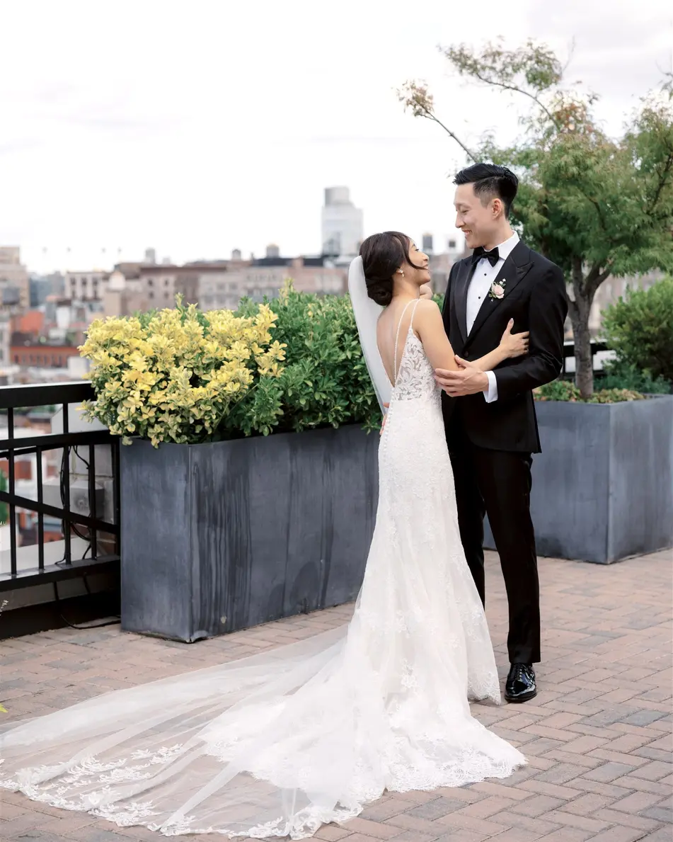 On a brick terrace edged with planters, the bride looks up at the groom, her long veil and train spread behind her.