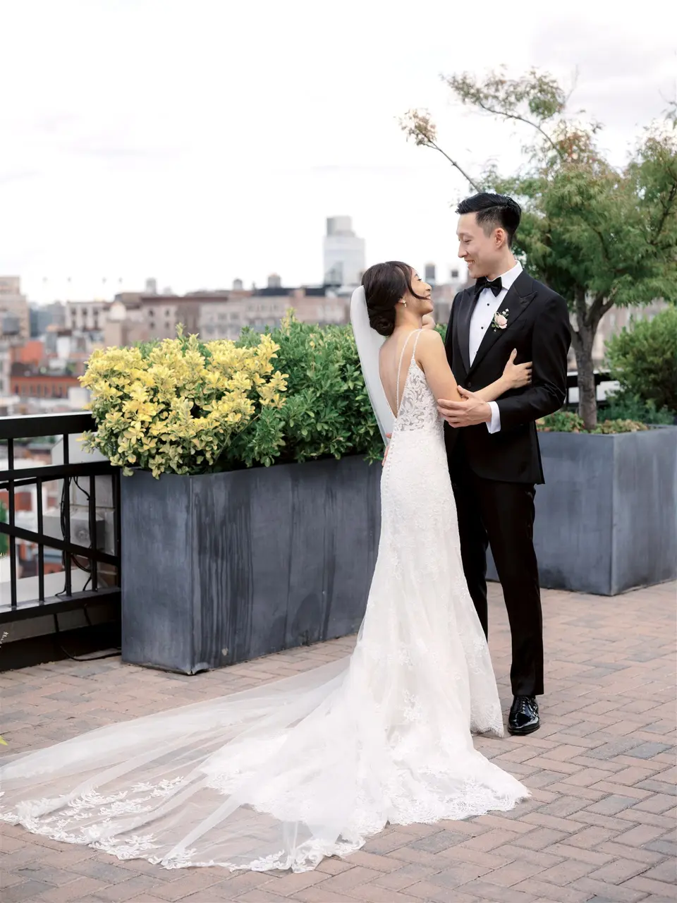 On a brick terrace edged with planters, the bride looks up at the groom, her long veil and train spread behind her.