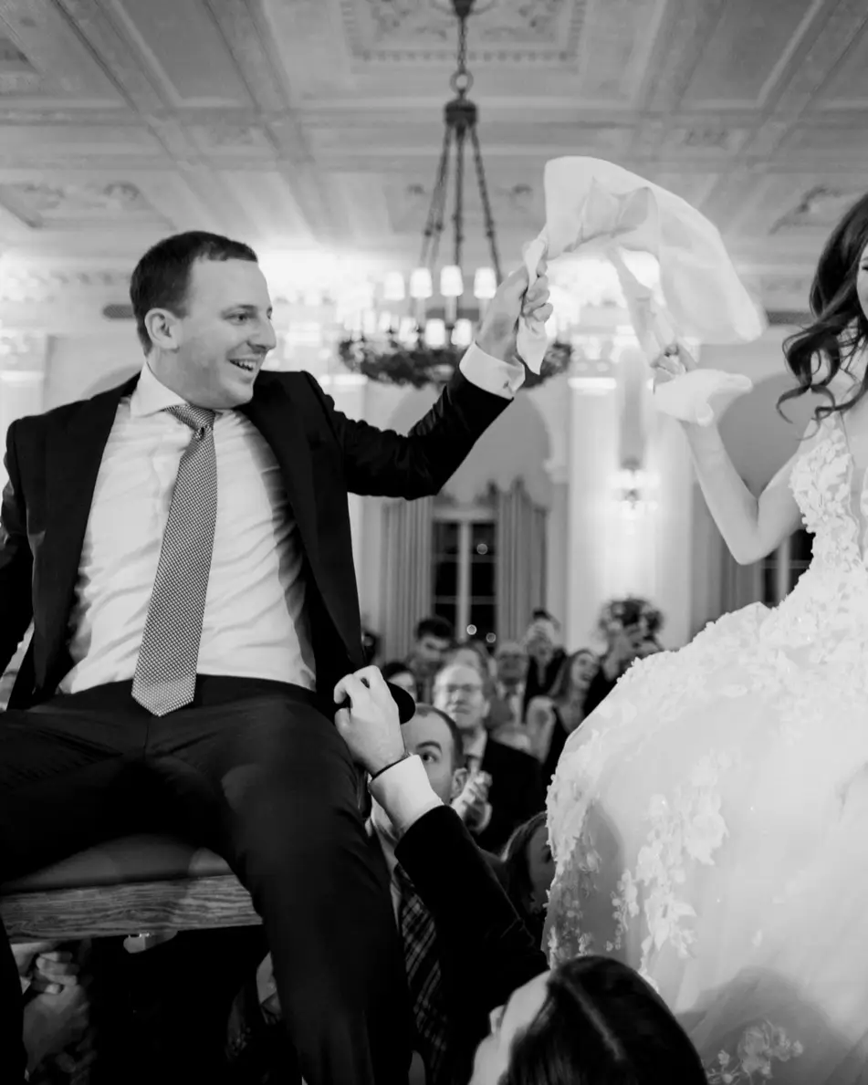 The couple are lifted high on chairs above a crowd of guests, the bride laughing and holding on. Black and white.