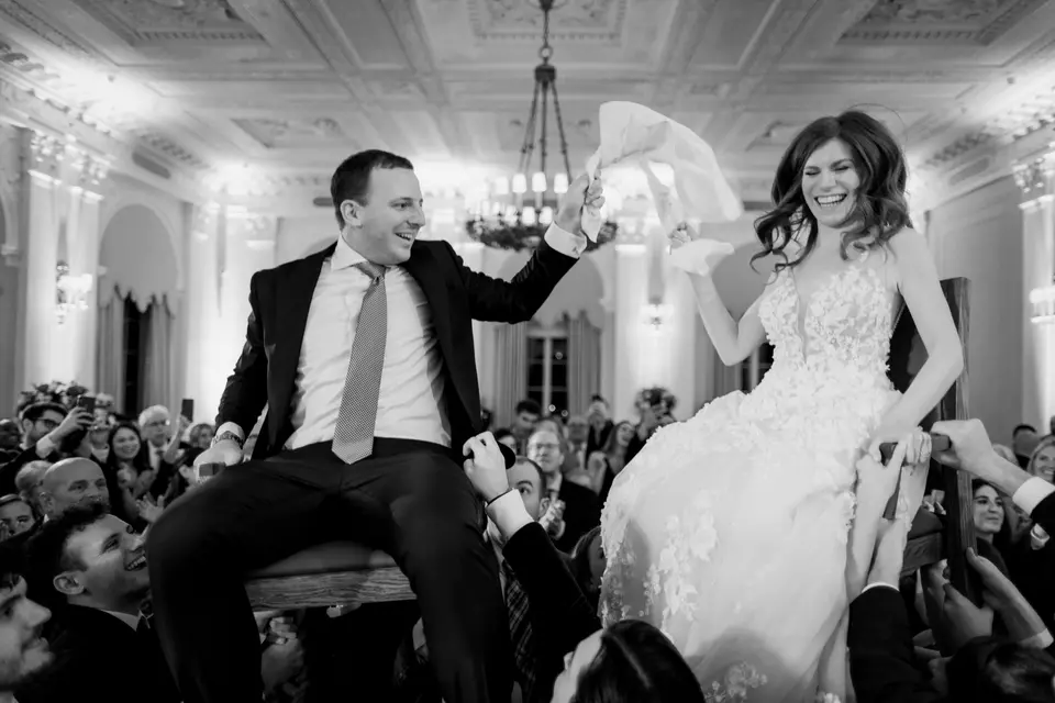 The couple are lifted high on chairs above a crowd of guests, the bride laughing and holding on. Black and white.