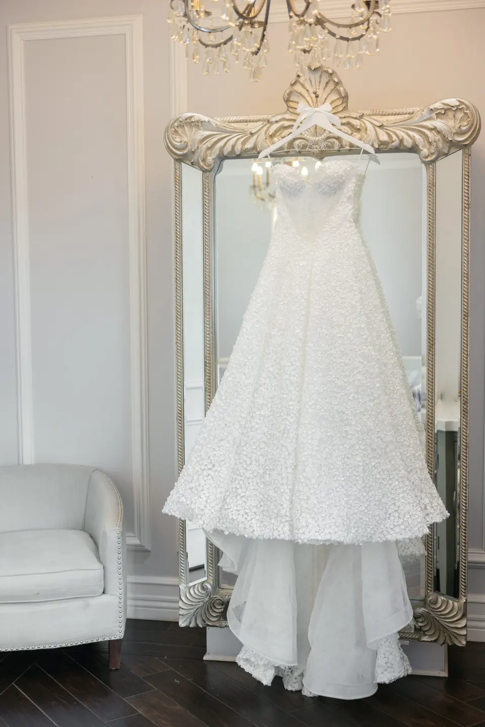 The wedding gown hangs from an ornate silver mirror in a white room, its tiered skirt falling to the floor.