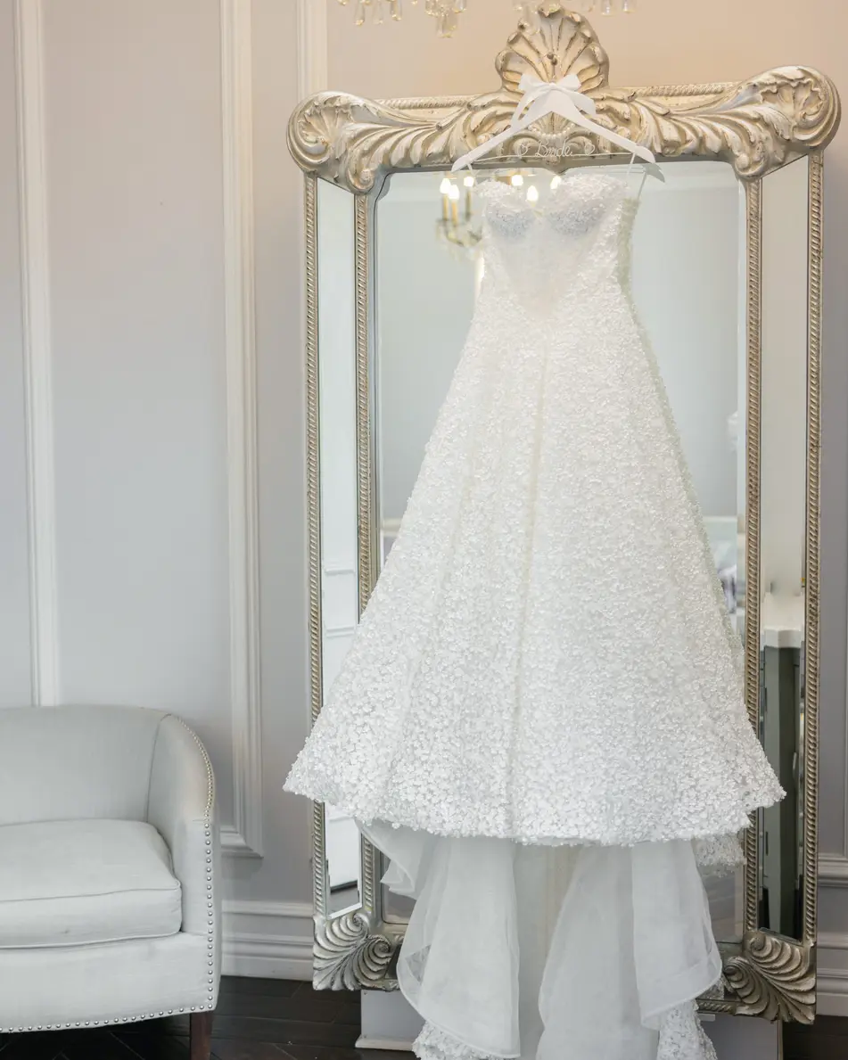 The wedding gown hangs from an ornate silver mirror in a white room, its tiered skirt falling to the floor.