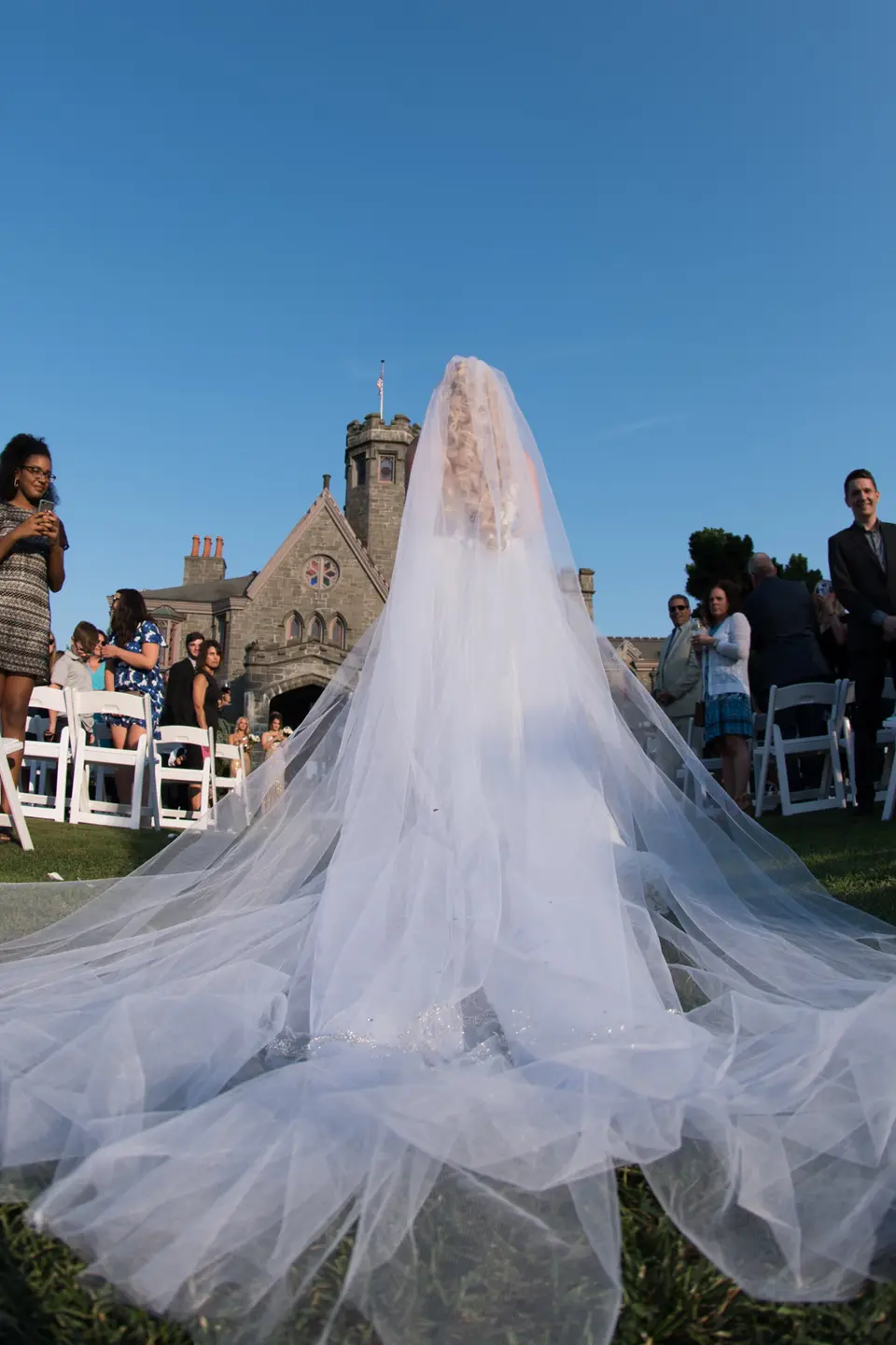 From behind, the bride's long veil spreads across the grass as she stands before the castle.