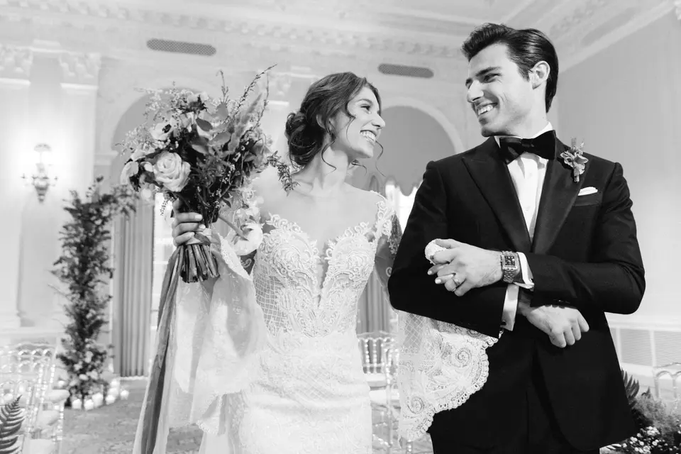 The bride and groom walk arm in arm, laughing, her bouquet raised. Black and white.