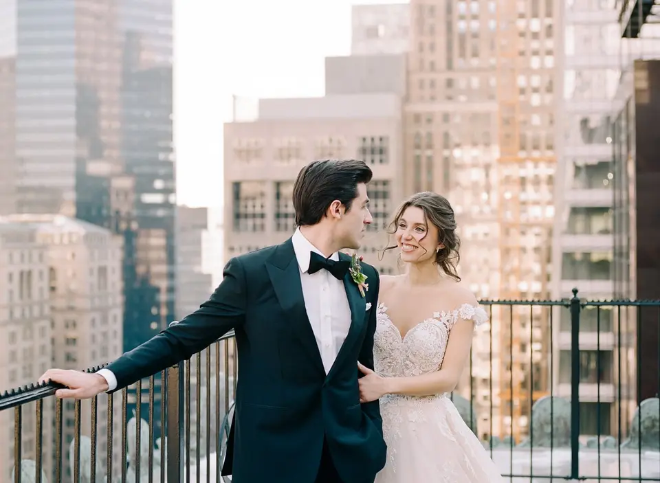 The groom turns at the railing to see the bride, smiling beside him on the terrace.