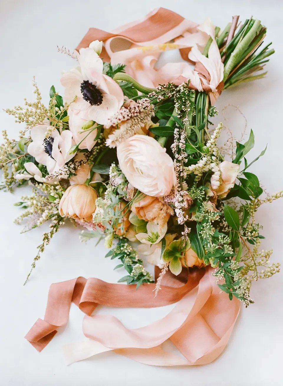 The bridal bouquet of blush anemones, ranunculus and greenery tied with long peach silk ribbons.
