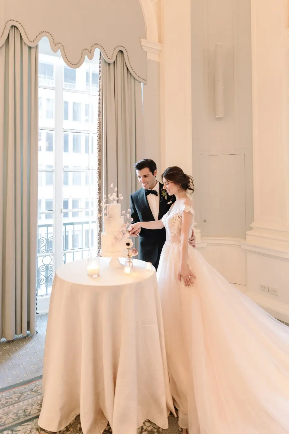 The bride and groom cut a small white cake by a tall window.