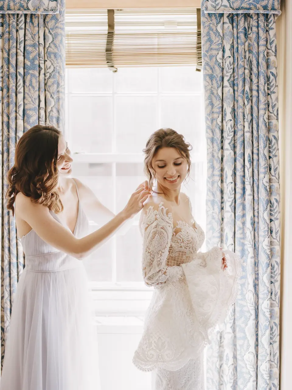 A bridesmaid helps the bride into her lace gown by a window hung with blue patterned curtains.
