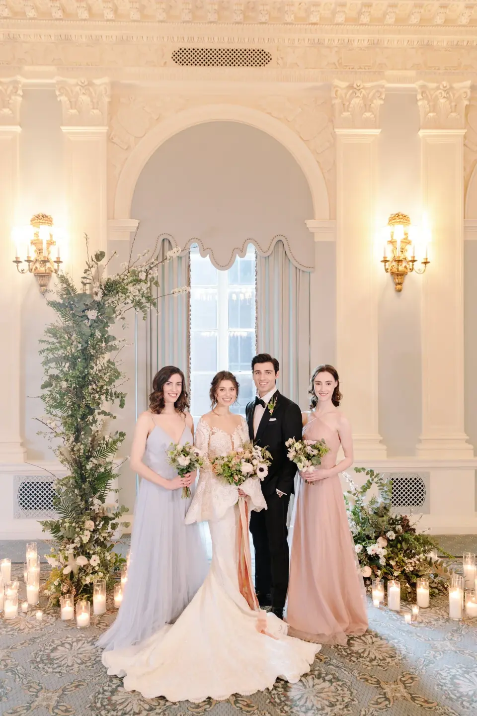 The couple and two bridesmaids beneath the column of flowers, candles on the carpet at their feet.