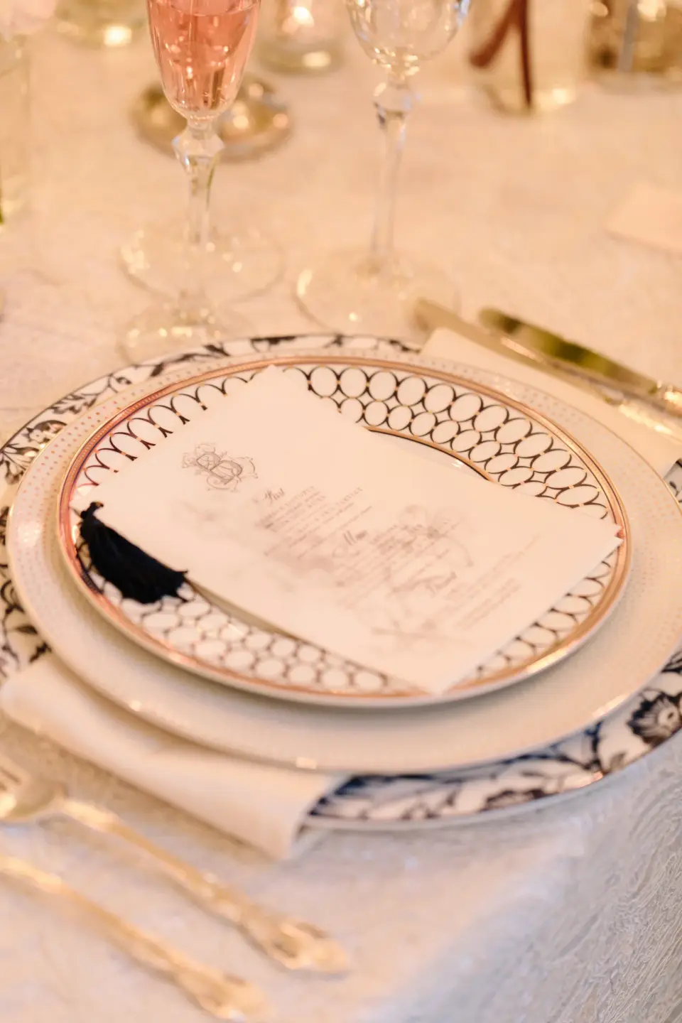 A menu card with a blue tassel on a patterned charger, a glass of rosé beside it.