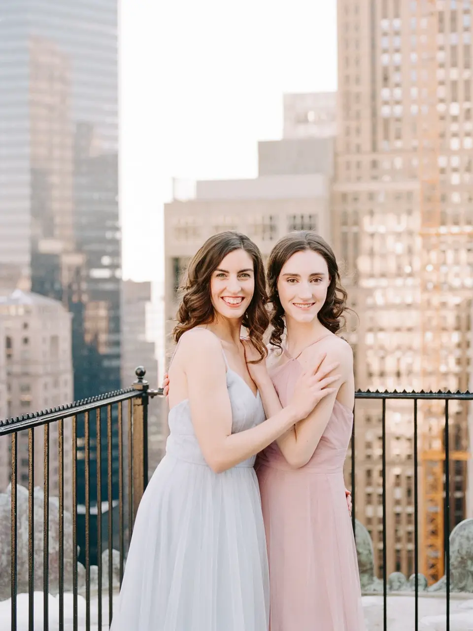 Two bridesmaids in blue and blush hug on the roof terrace, Midtown behind them.
