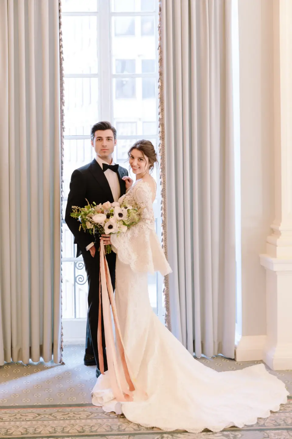 The bride and groom by a tall window, her bouquet ribbons trailing to the floor.