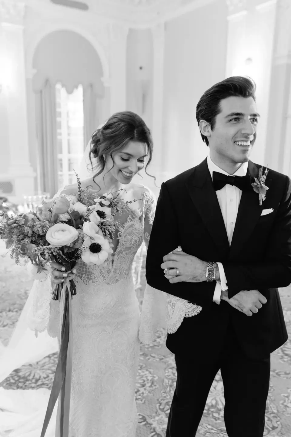 The bride and groom walk arm in arm, smiling. Black and white.